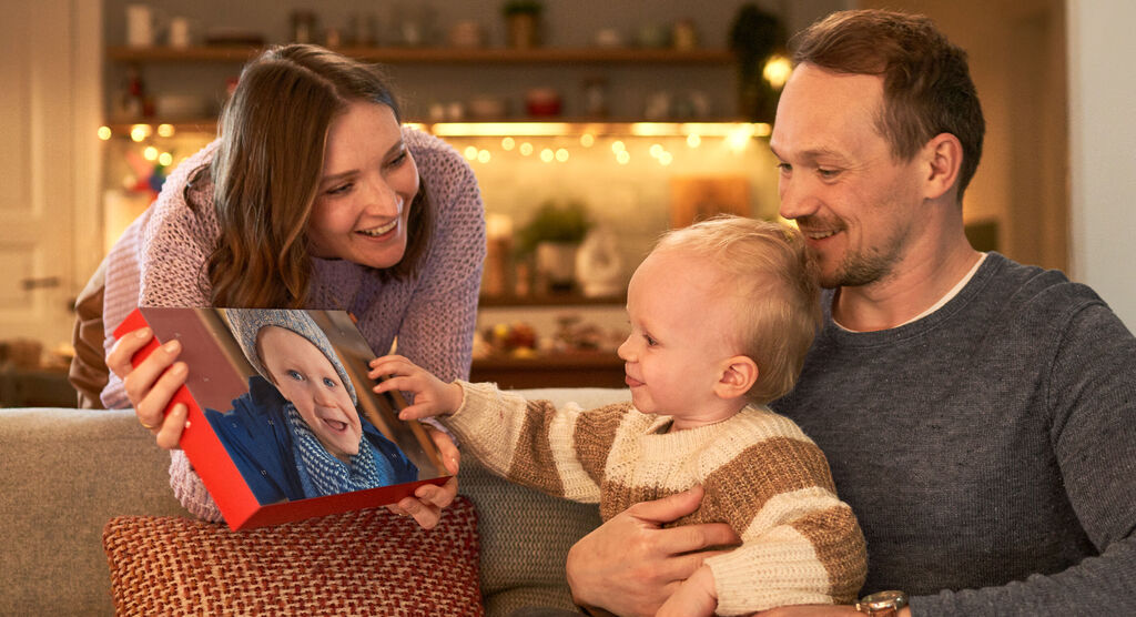 Rodina sedí v útulnej obývačke a pozerá si fotoknihu. Dieťa ukazuje na knihu a usmieva sa, zatiaľ čo dospelí pozerajú nadšene.