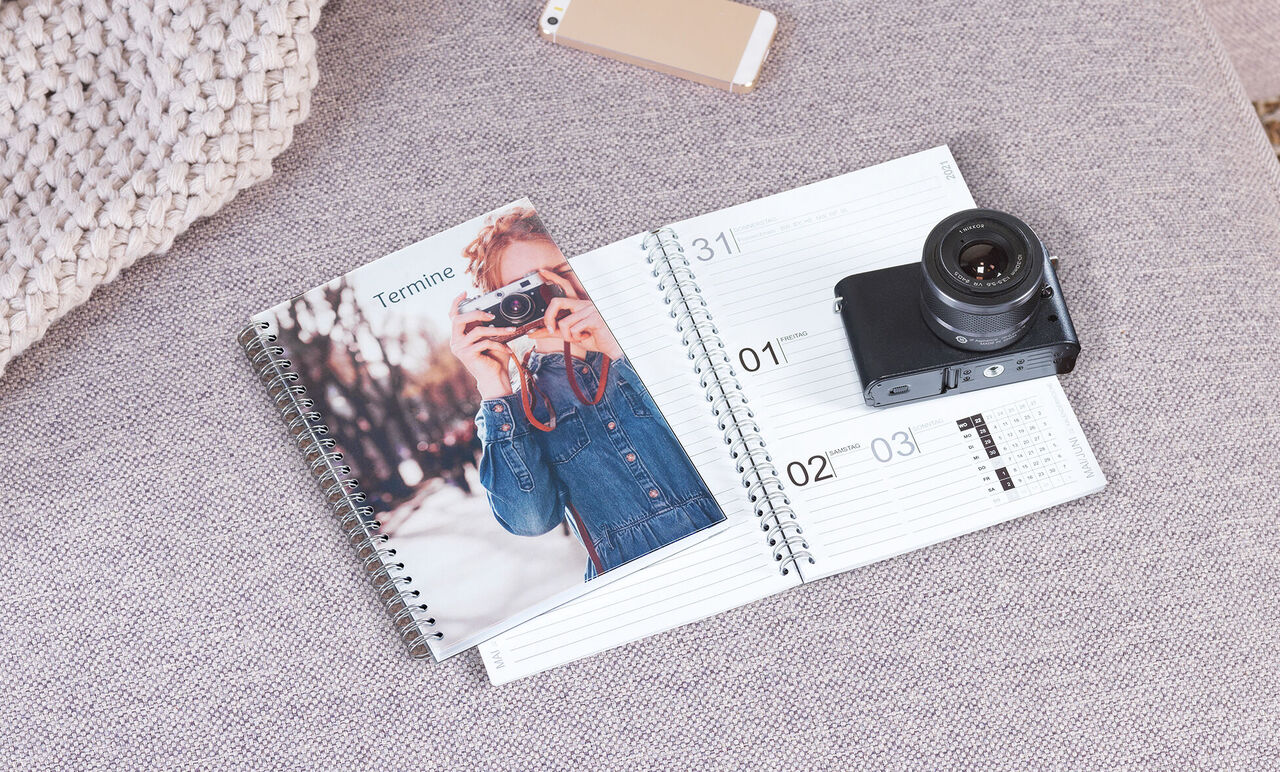 Otvorený zápisník s fotoaparátom a fotoknihou na sivom gauči.