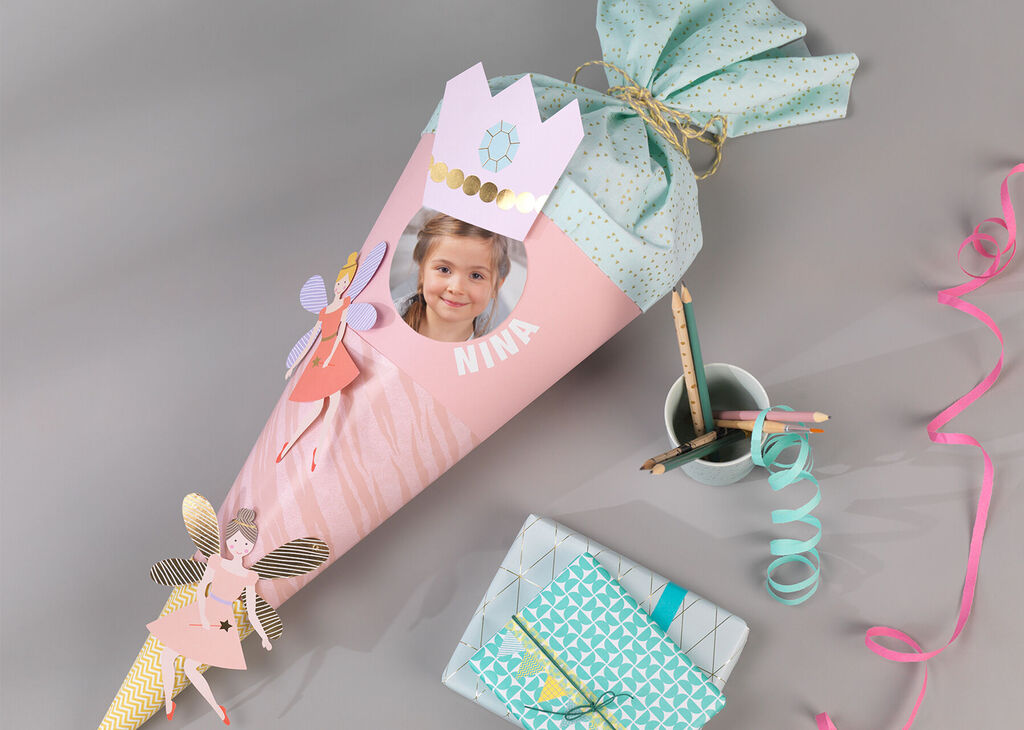Farebný školský kužeľ s princeznou a vílou, ozdobený zlatou stuhou a farebným papierom, na sivom stole.
