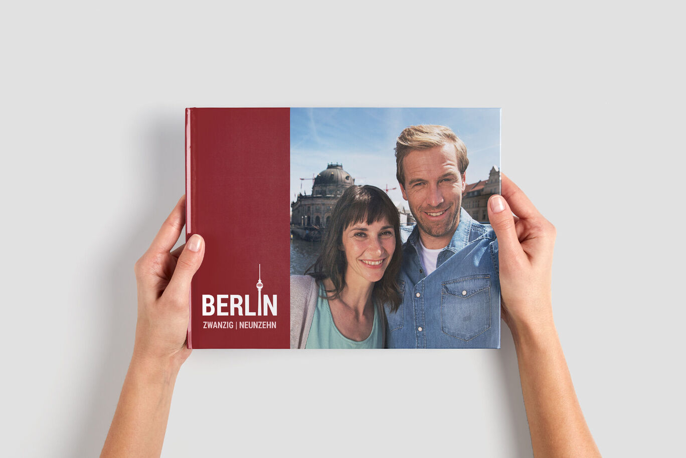Dve osoby držia CEWE fotoknihu s názvom 'Berlín', na sivom pozadí.