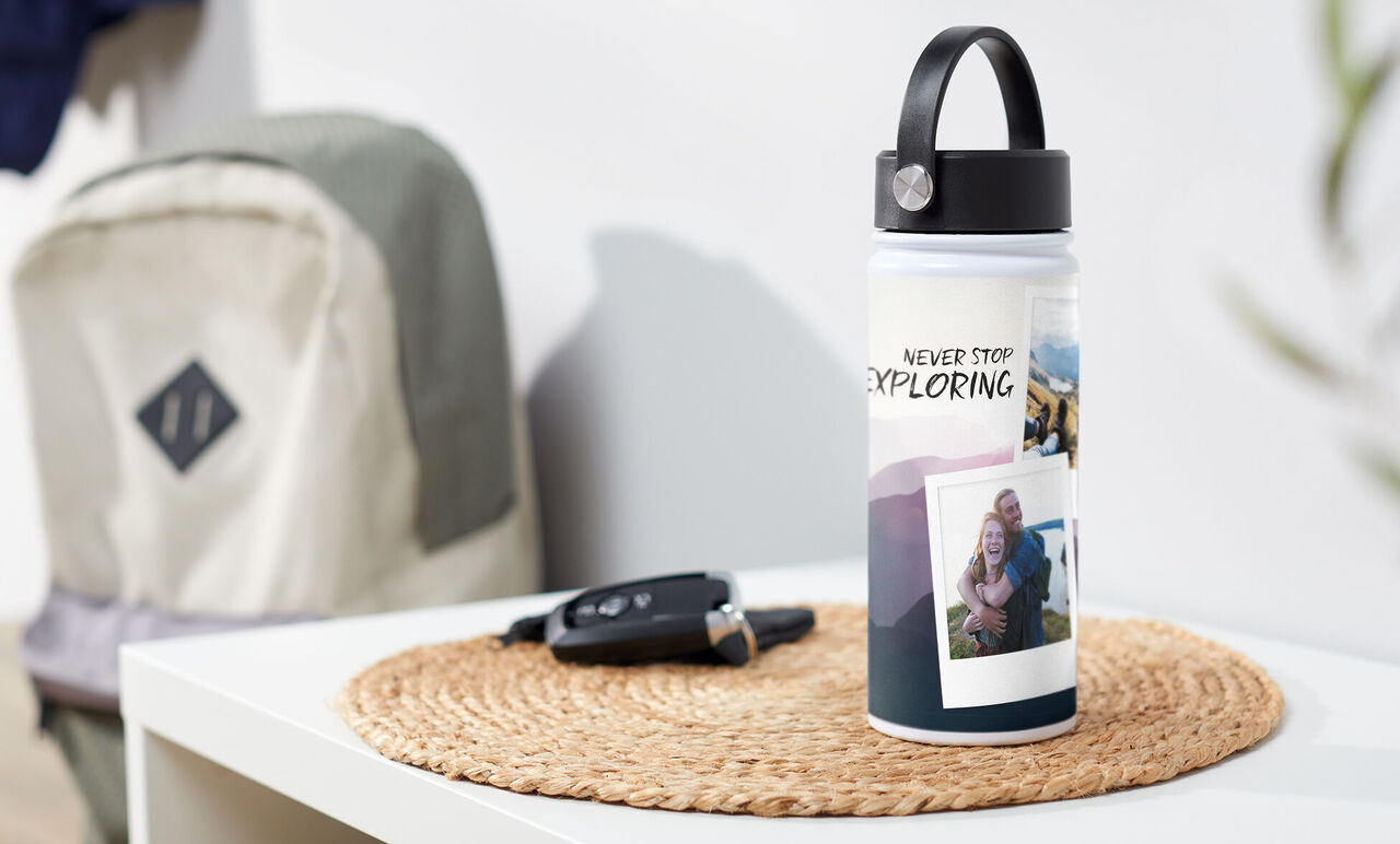 Fľaša s nápisom „Never Stop Exploring“ a fotografiami na stole. V pozadí batoh a kľúče od auta.