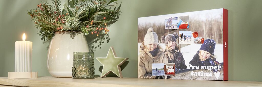 CEWE fotokniha stojí na stole, obklopená sviečkou, zelenou hviezdou a rastlinou v kvetináči. Zobrazuje zimnú fotokoláž s tromi usmievajúcimi sa deťmi v teplom oblečení vonku.