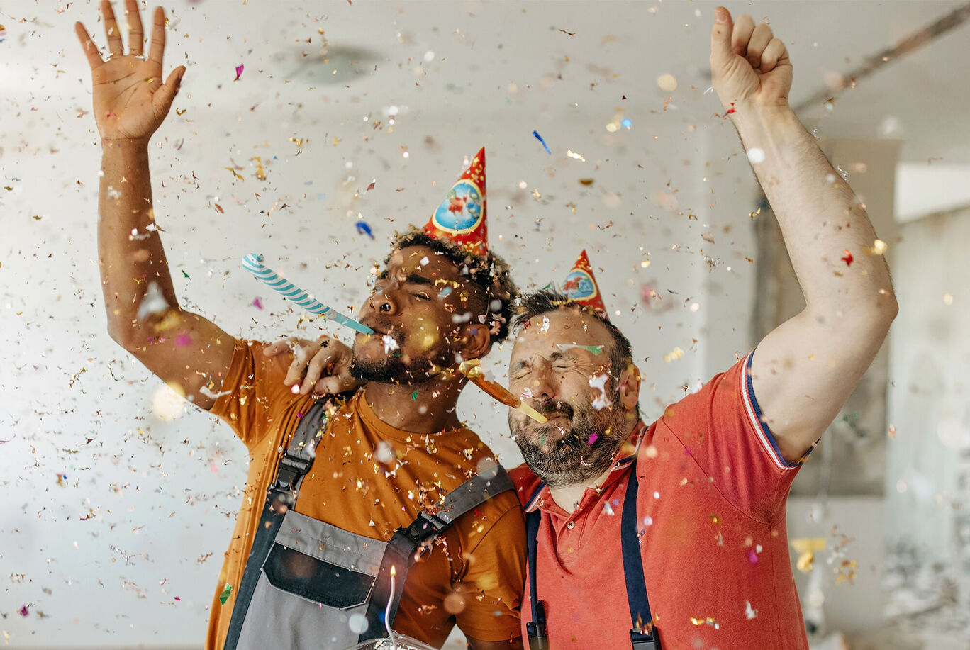 Dvaja oslavujúci ľudia s party klobúkmi v pestrofarebnom prostredí, obklopení konfietami.