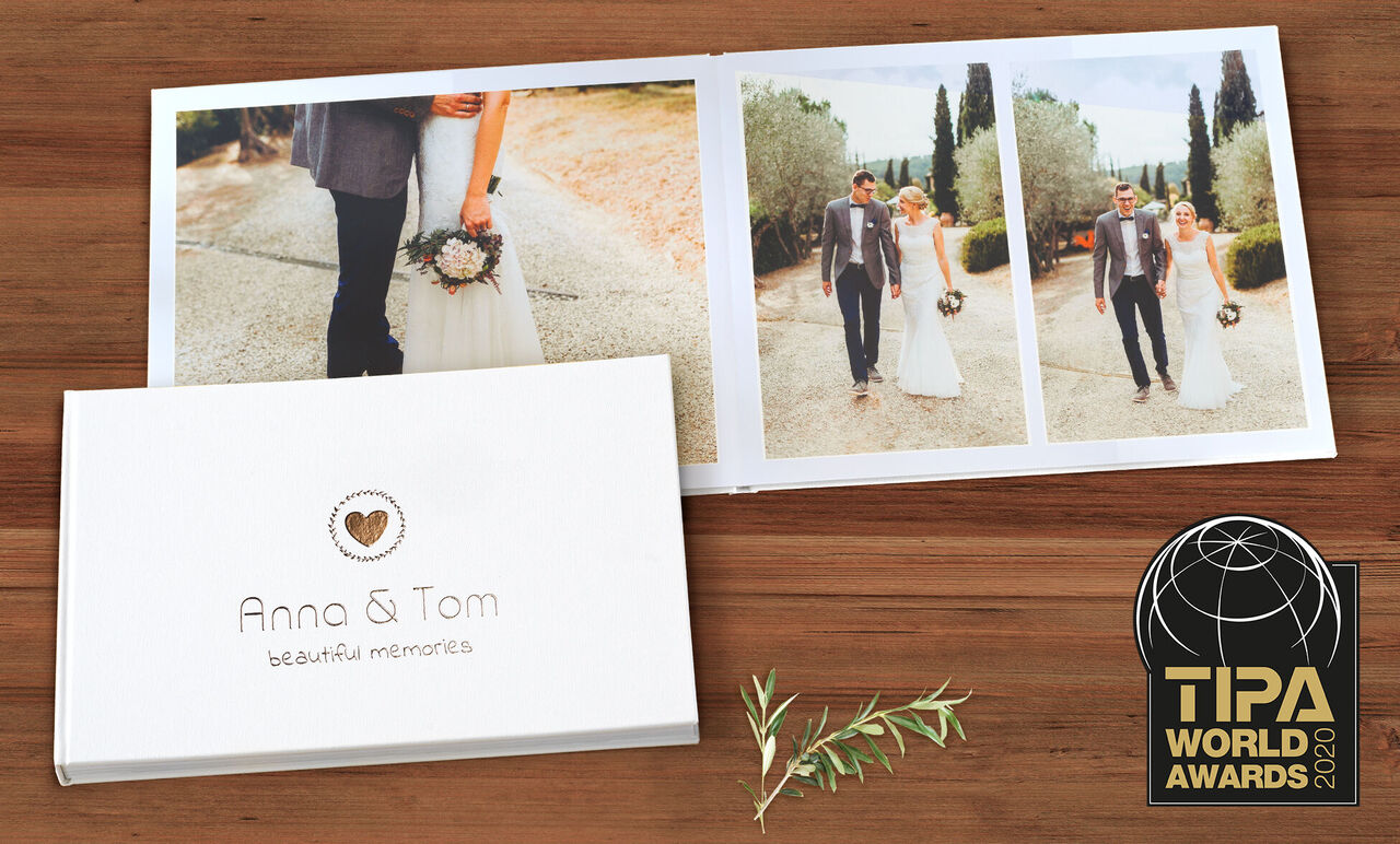 CEWE FOTOKNIHA s názvom "Anna & Tom - krásne spomienky" leží otvorená na drevenom stole. Vľavo je biely dizajn obalu, vpravo sú fotky páru vo svadobnom oblečení.
