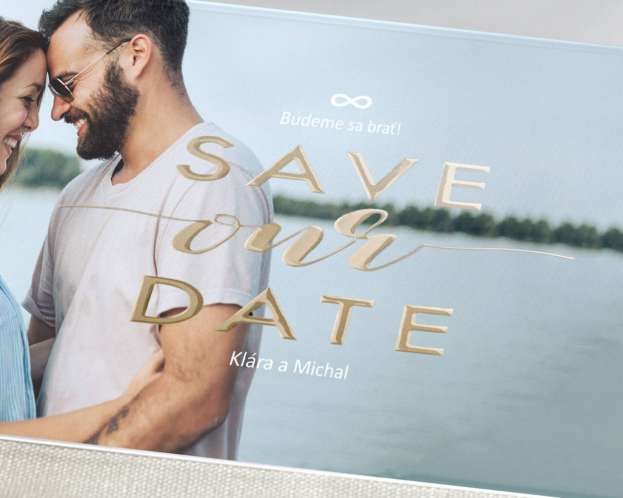 Pár stojí pri vode a drží tabuľku Save-the-Date.