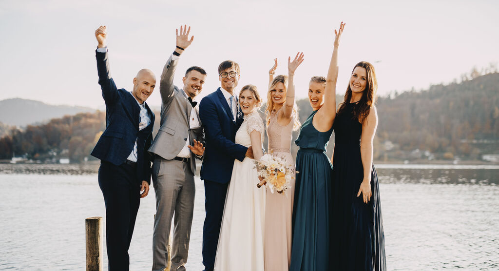 Priatelia oslavujú pri jazere, elegantne oblečení, s úsmevmi a mávaním.