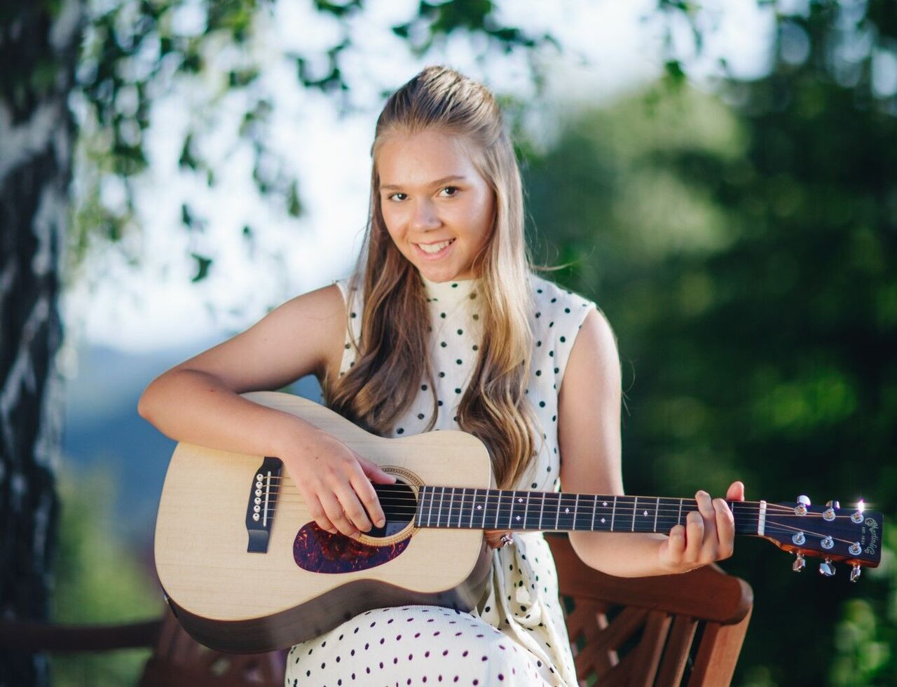 Žena hrá na gitare vonku pred zelenou scenériou.