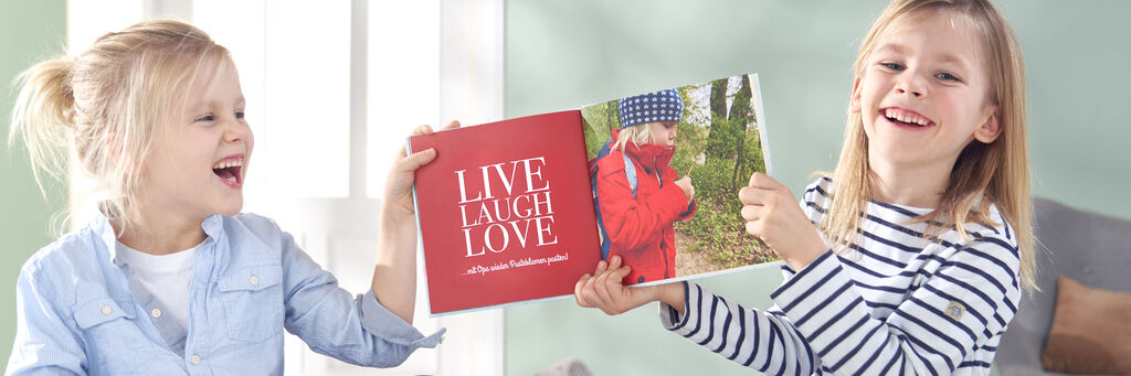 Dve deti držia otvorenú fotoknihu a usmievajú sa. Fotokniha má nápis "LIVE LAUGH LOVE".