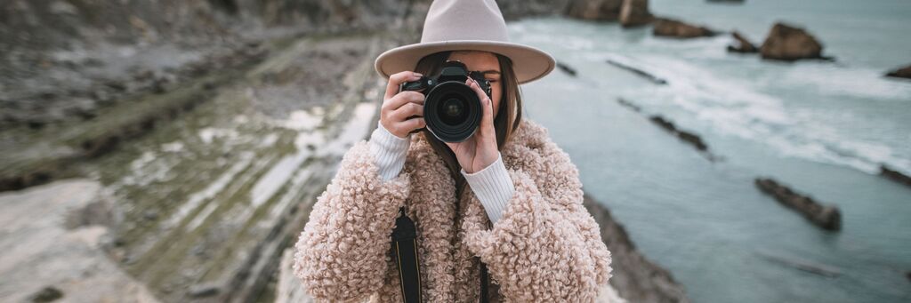 Osoba drží fotoaparát a usmieva sa na pobreží, obklopená skalami.