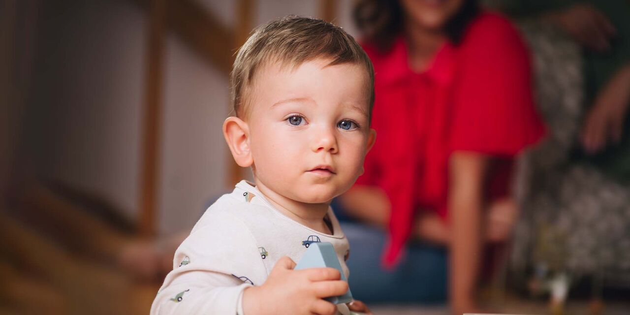 Malé dieťa drží hračku a usmieva sa do kamery. V pozadí si dospelí užívajú čas.