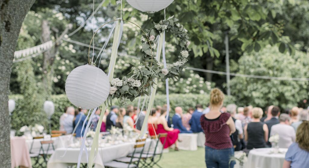 Svadobná oslava v záhrade s dekoratívnymi lampášmi a kvetinovým vencom.