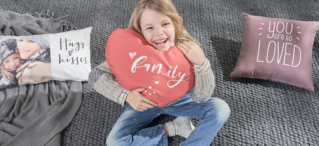 Dieťa sedí na koberci s dvomi dekoračnými vankúšmi. Jeden je v tvare srdca s nápisom 'family' a druhý má text 'You are so LOVED'.