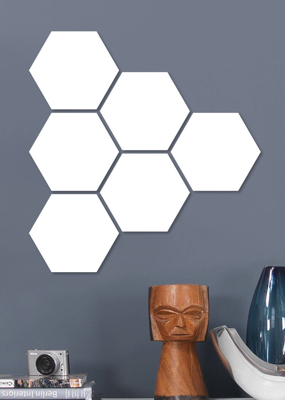 Šesť bielych hexagónov na šedej stene vedľa modernej lampy a fotoaparátu.