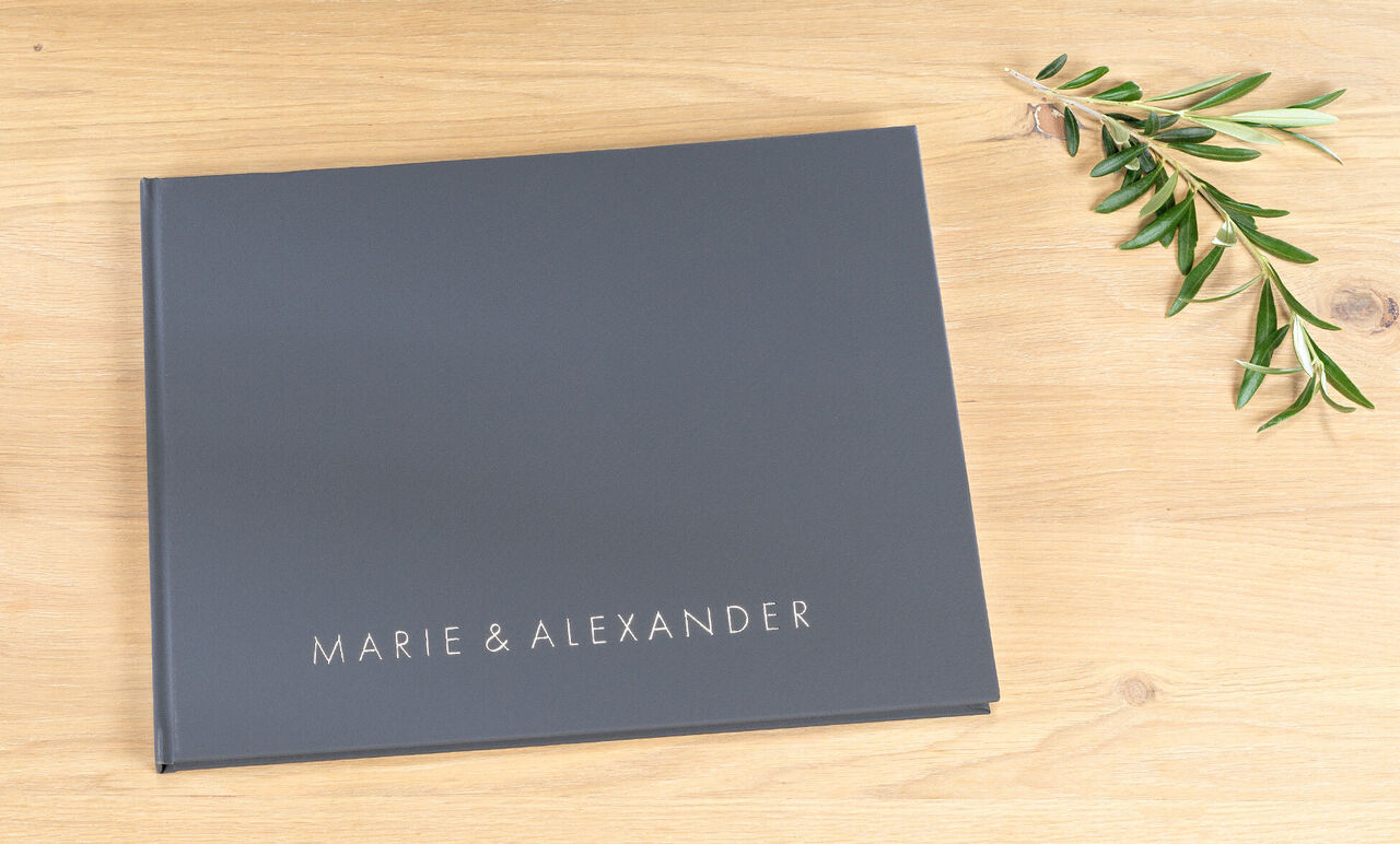 Tmavá fotokniha s názvom „Marie & Alexander“ na drevenom povrchu s olivovou vetvičkou