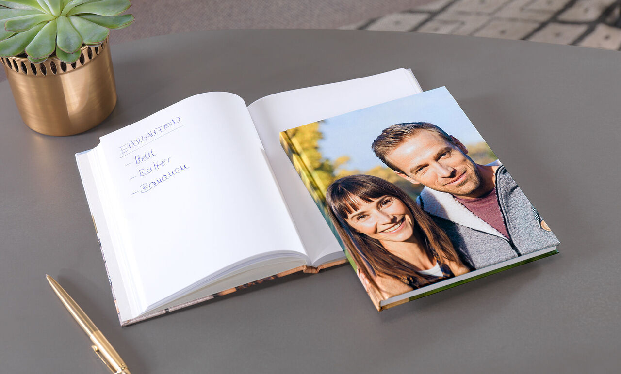 Otvorená fotokniha a kalendár na stole, obklopené zelenými rastlinami.