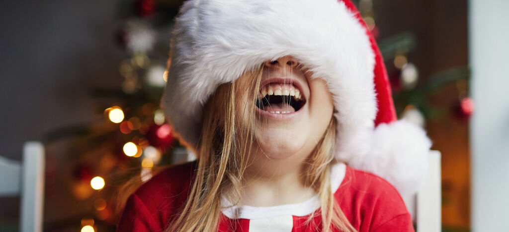 Dieťa má na sebe čiapku Santa Clausa a sedí pred vianočným stromčekom.