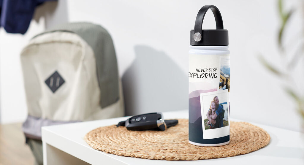 Personalizovaná fľaša s fotkami na stole vedľa kľúčov a batohu s textom "NEVER STOP EXPLORING".