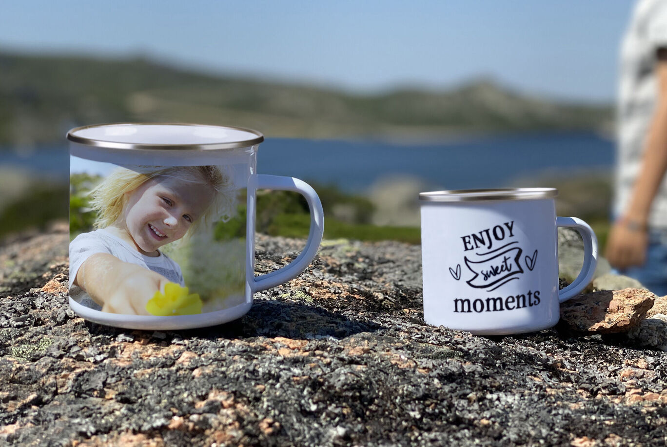 Dve šálky na skale s pekným výhľadom v pozadí. Jedna má fotku usmievajúceho sa dieťaťa, druhá nápis 'ENJOY sweet moments'.