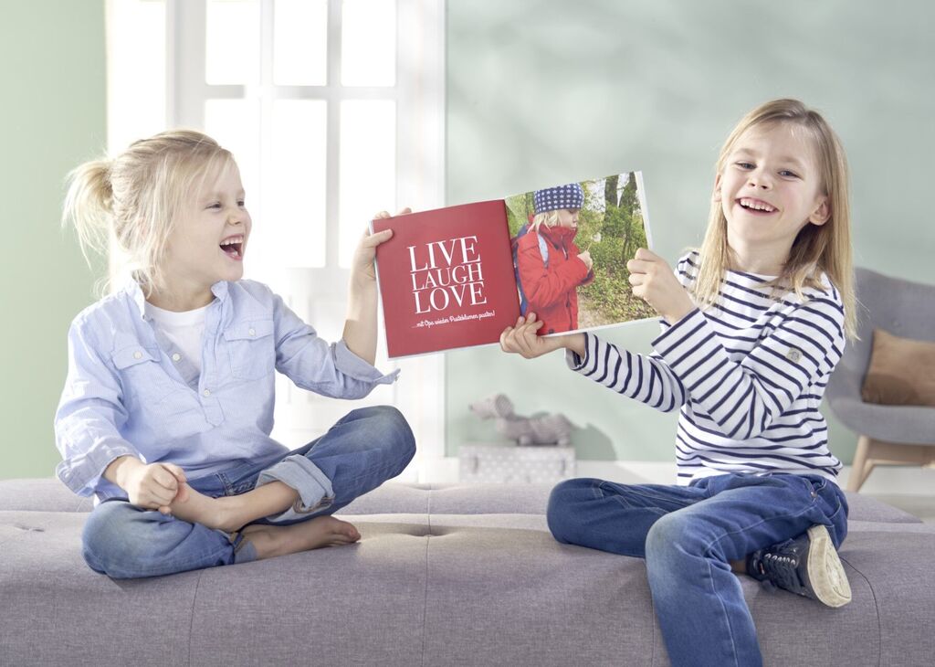 Dvaja deti sedia na pohovke a ukazujú krásne CEWE fotoknihu. Obal nesie text „LIVE LAUGH LOVE“ a farebné obrázky.