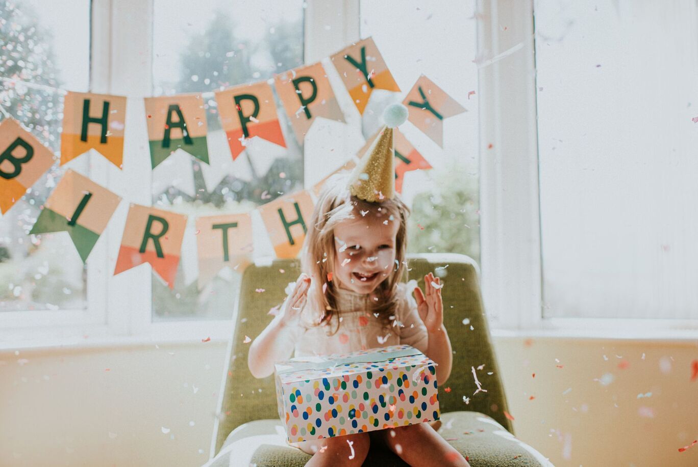 Šťastné dieťa na stoličke drží farebne zabalený darček. Za ním je narodeninový banner s nápisom "HAPPY BIRTHDAY".