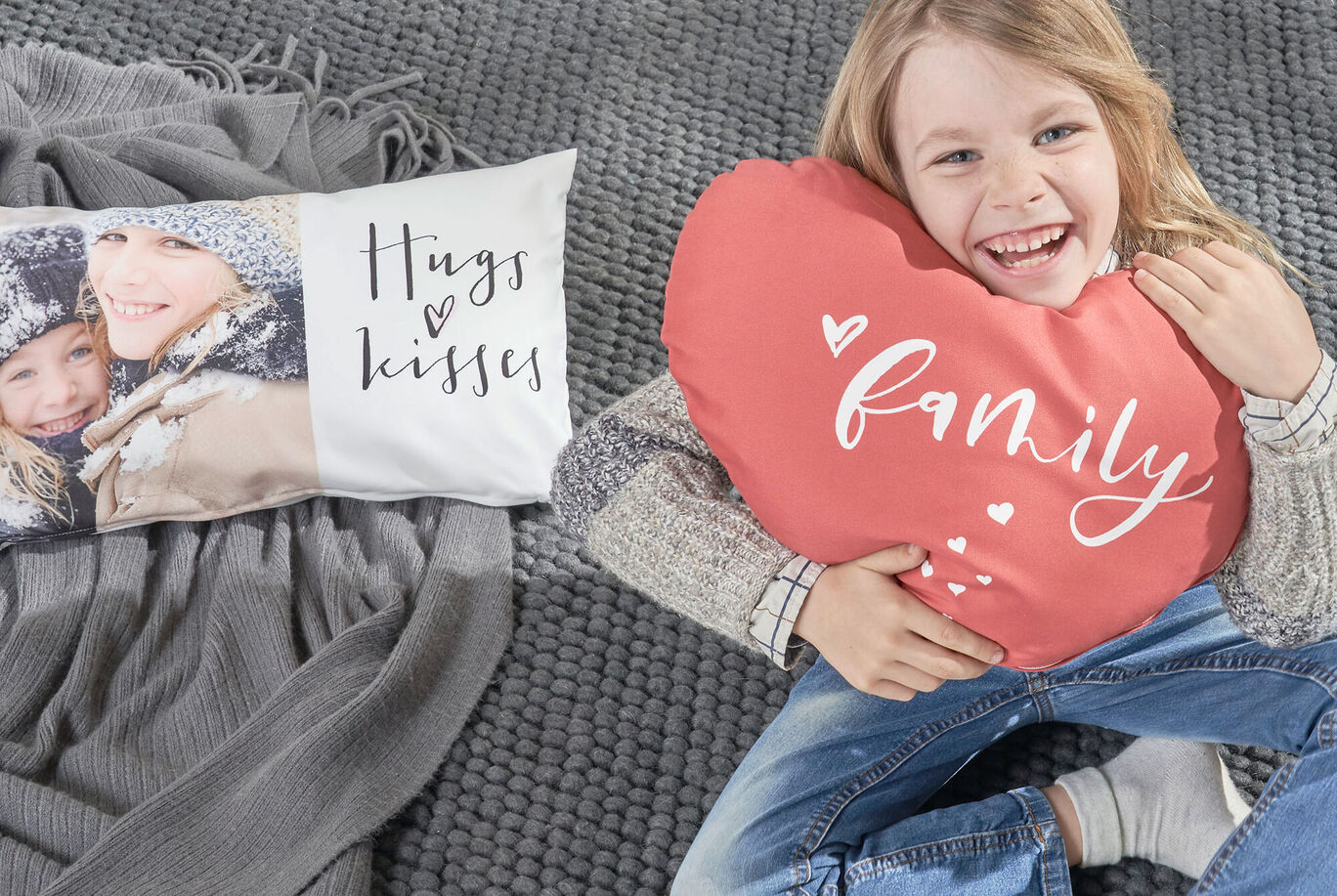 Osoba sedí na koberci a drží vankúš v tvare srdca s nápisom „family“. Naľavo je vankúš s textom „Hugs & Kisses“.
