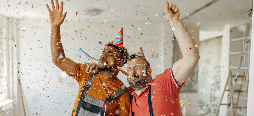 Dvaja veselí muži oslavujú s konfietami a party klobúkmi v jasnej miestnosti.