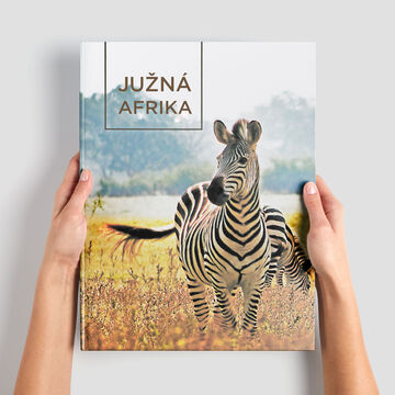 Osoba drží CEWE FOTOKNIHU s názvom "Južná Afrika" pred krásnou krajinou so zeberami.