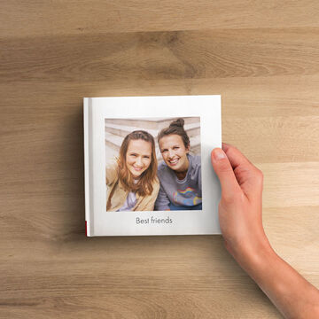 CEWE FOTOKNIHA s nápisom 'Best friends' na drevenom stole, ruka drží knihu.