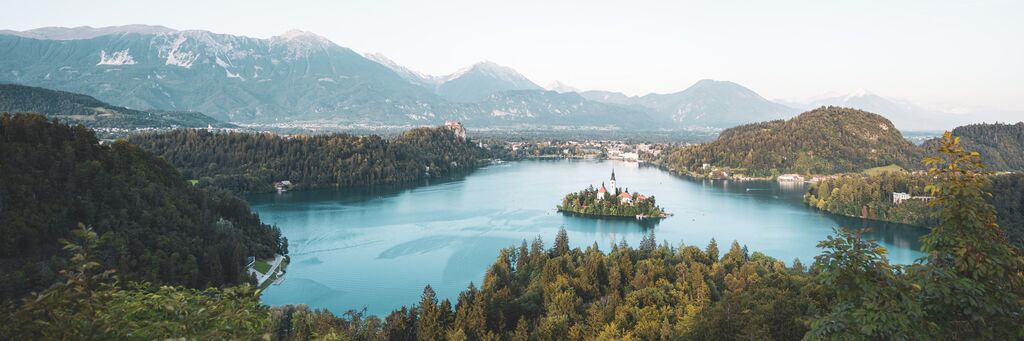Panoramatický výhľad na Bledské jazero s ostrovom a hradom v popredí, obklopené horami a lesmi v jemných farbách.