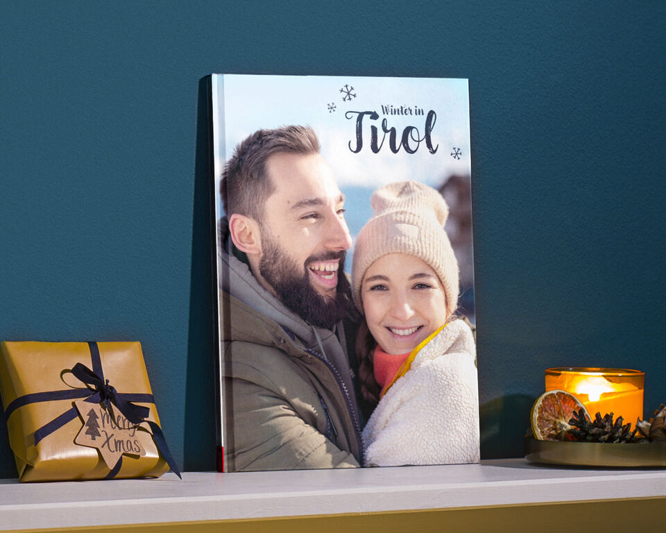 CEWE FOTOKNIHA so slovami „Zima v Tirolsku“ na stole vedľa darčeka a sviečky