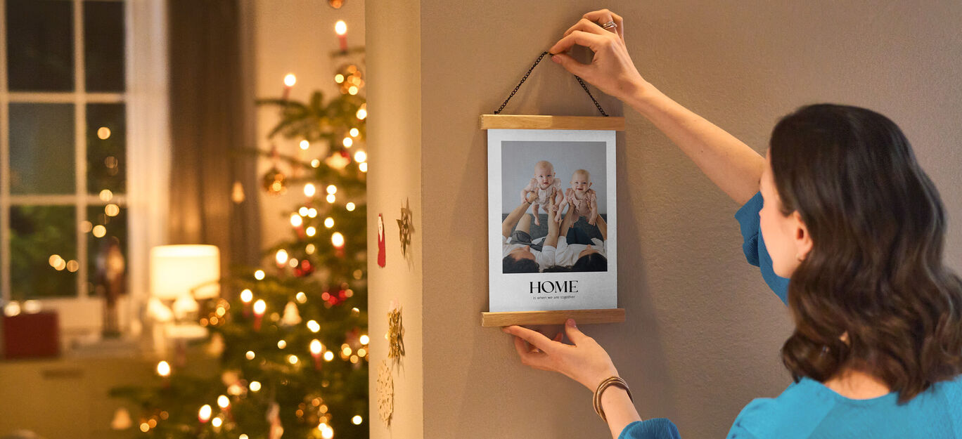 Osoba umiestňuje zarámovaný obraz s nápisom "HOME" na stenu. V pozadí stojí osvetlený vianočný stromček.