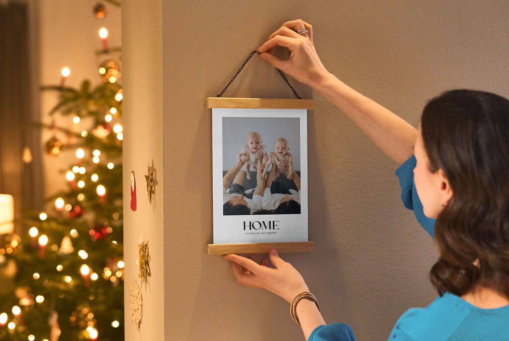 Osoba vešia zarámovanú fotografiu s nápisom "HOME" na stenu. V pozadí svieti vianočný stromček.