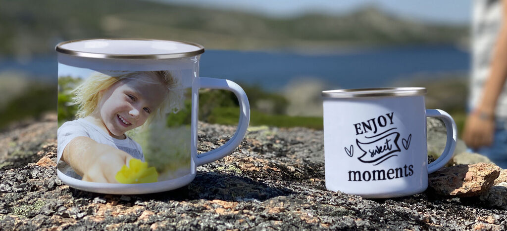 Dve šálky na kameni s výhľadom na krajinu. Jedna má veselý obrázok dieťaťa s kvetinou, druhá text: ENJOY sweet moments.