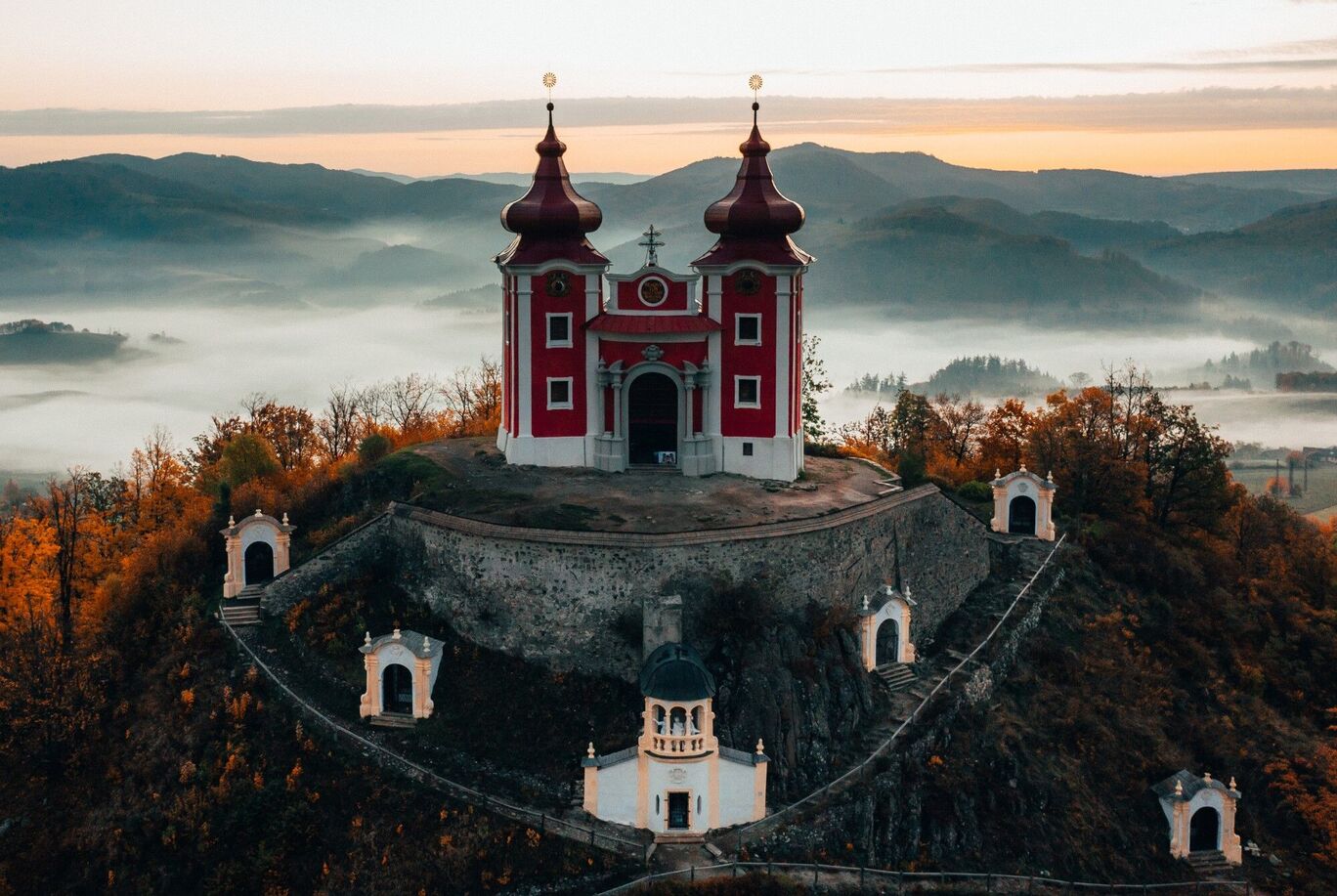Červený kostol na kopci obklopený jesennými stromami a hmlou v pozadí