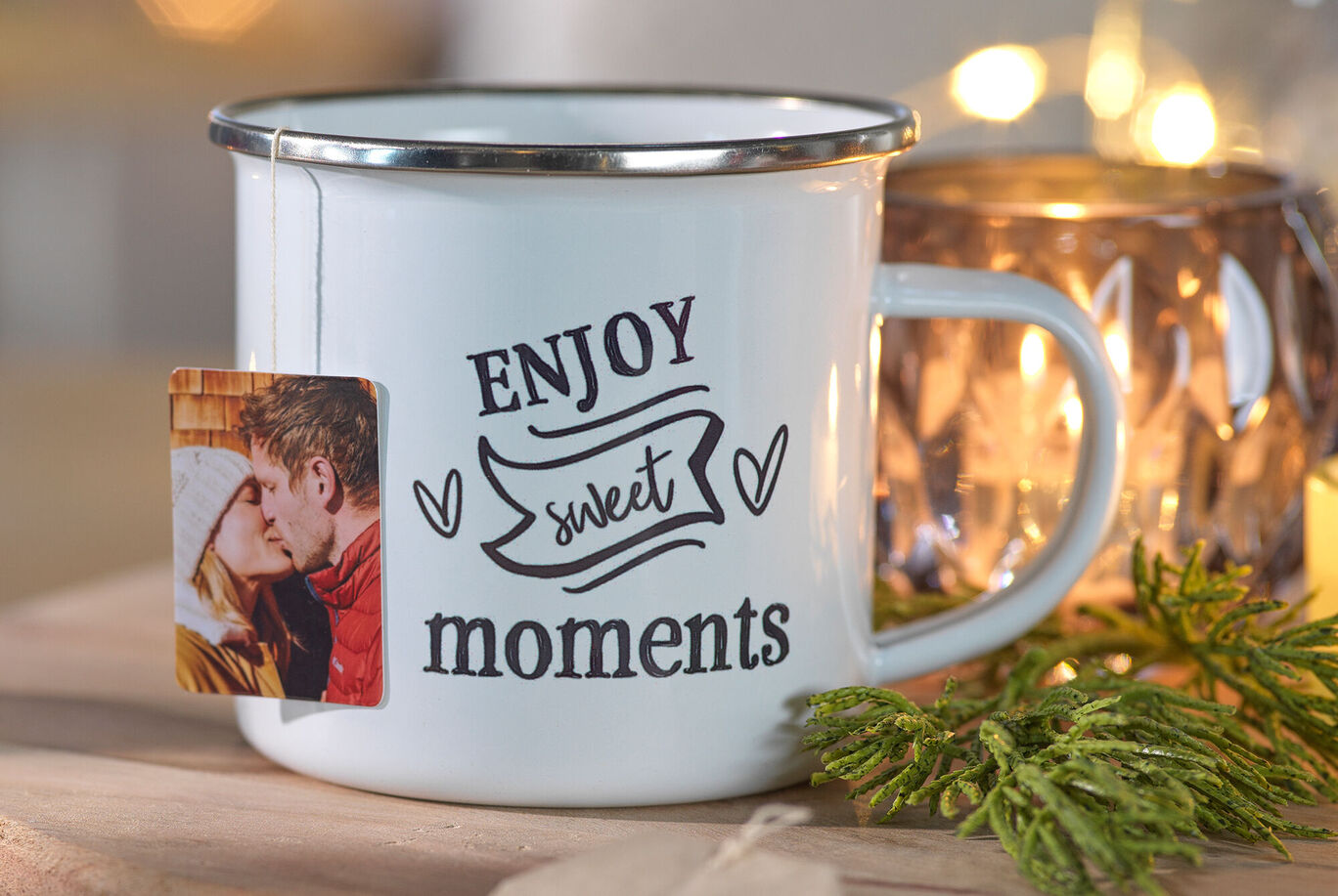 Biely hrnček s nápisom „Enjoy sweet moments“. Na ňom visí fotoprívesok, vedľa je čajová sviečka a vetvičky.